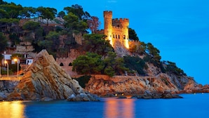 A coastal castle with a stone tower, surrounded by rocky terrain and trees, overlooking a calm sea. - Hotel Boutique Can Colom (Lloret de Mar)