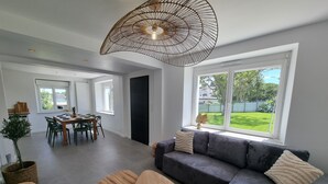 Una sala moderna con un sofá gris, dos mesitas de madera, una zona de comedor con mesa y sillas, y un amplio ventanal con vistas al jardín.
