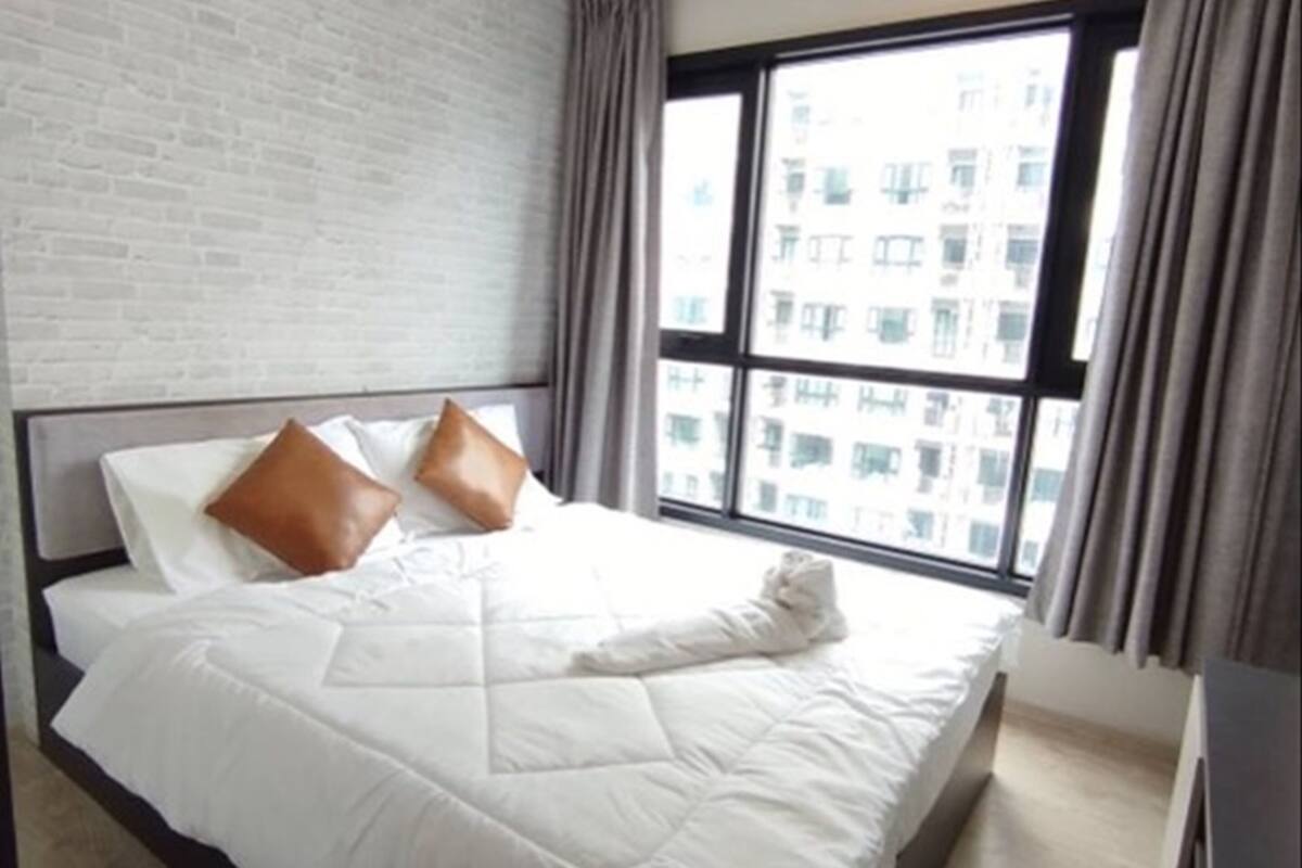 Ein ordentlich gemachtes Bett mit weißer Bettwäsche und zwei braunen Kissen, eine Backsteinwand und ein Fenster mit Vorhängen.