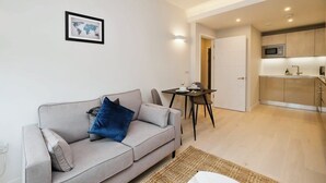 A modern living room with a grey sofa, a small table, and a kitchen area visible through an open door.