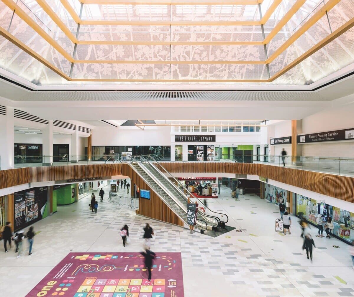 A multi-level shopping mall with escalators, stores, and a large open space with a patterned floor.