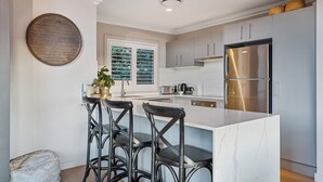 A modern kitchen with a central island, bar stools, stainless steel appliances, and a large window with blinds.