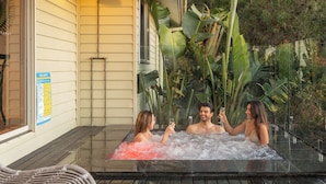 A hot tub with two people and a third person outside, surrounded by lush greenery.