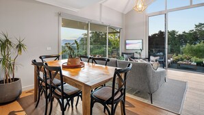 A dining area with a wooden table and chairs, a grey sofa, and a large window with a view of greenery and mountains.