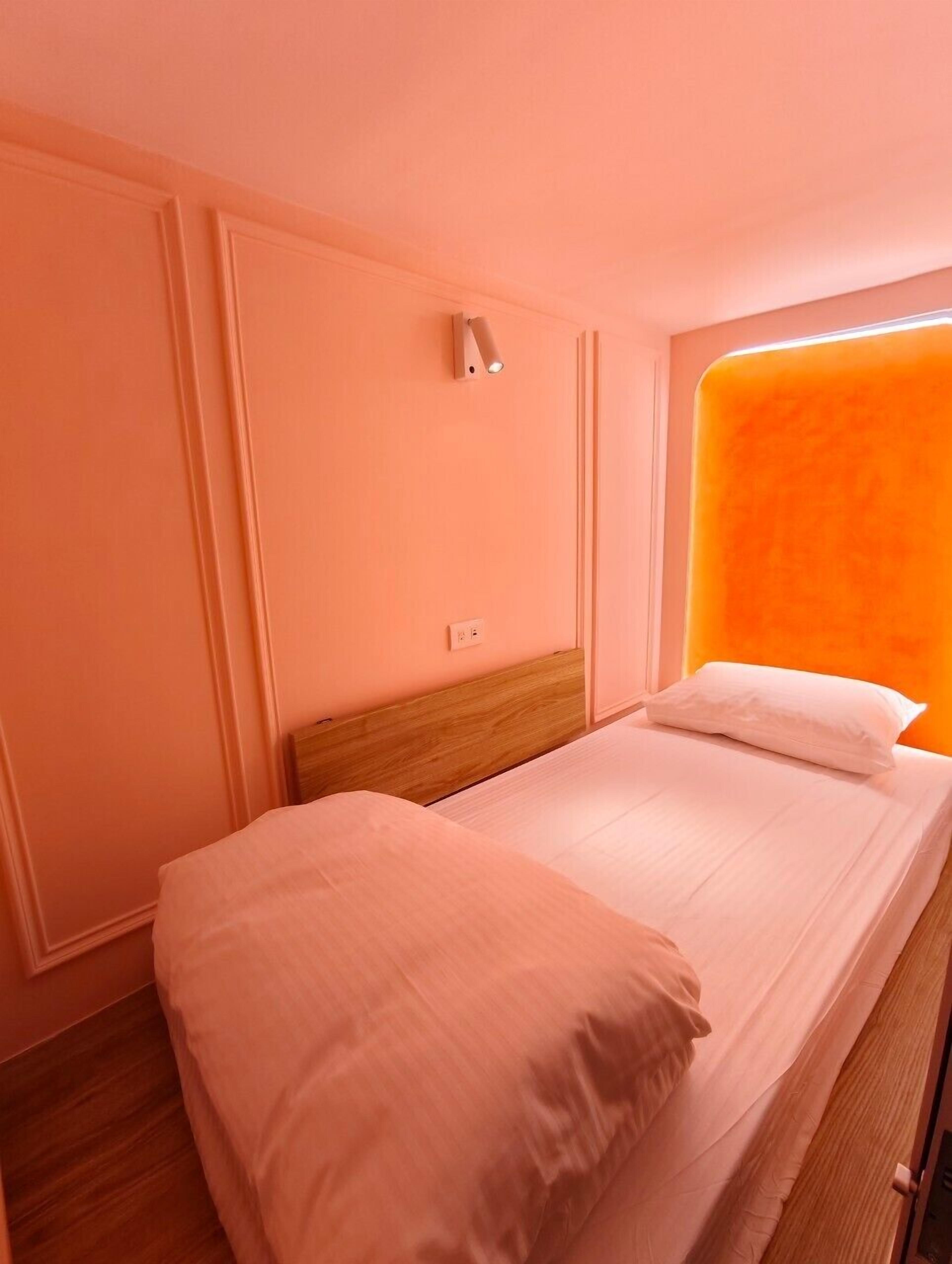 A single-bed room with a wooden headboard, a wall-mounted light, and a window with an orange accent.