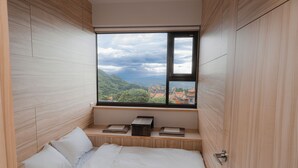A bedroom with a large window, wooden walls, a bed, and a small wooden shelf with books.