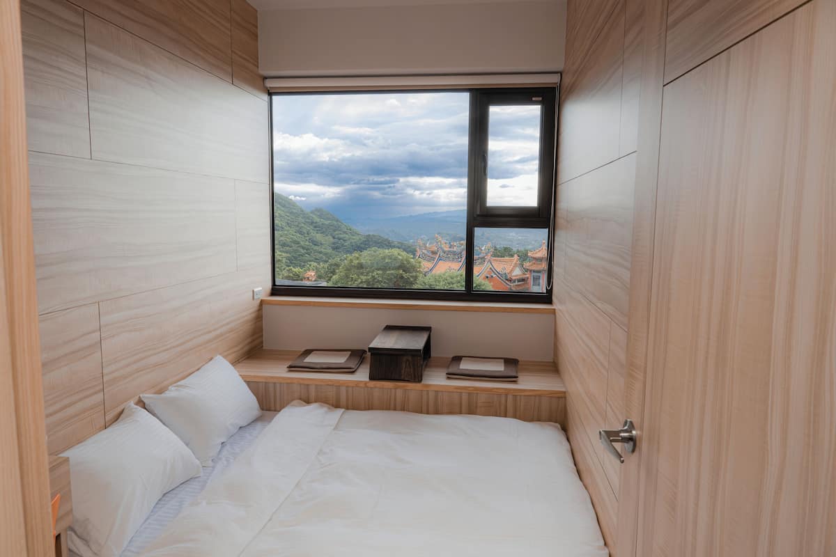 Een slaapkamer met een groot raam, houten wanden, een bed en een klein houten plankje met boeken.
