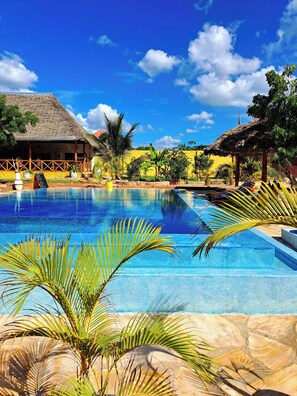 A clear blue pool with palm trees in the foreground, a thatched-roof building, and a yellow wall in the background. - Mimi na wewe villa (Nungwi)