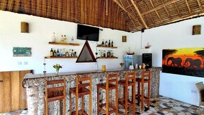 A bar area with a thatched roof, a TV mounted on the wall, and a variety of bottles on shelves. - Mimi na wewe villa (Nungwi)