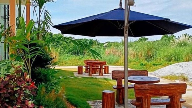 An outdoor seating area with wooden tables and chairs, surrounded by lush greenery and a stone pathway. - Jeanines Travellers Inn by RedDoorz (Morong)