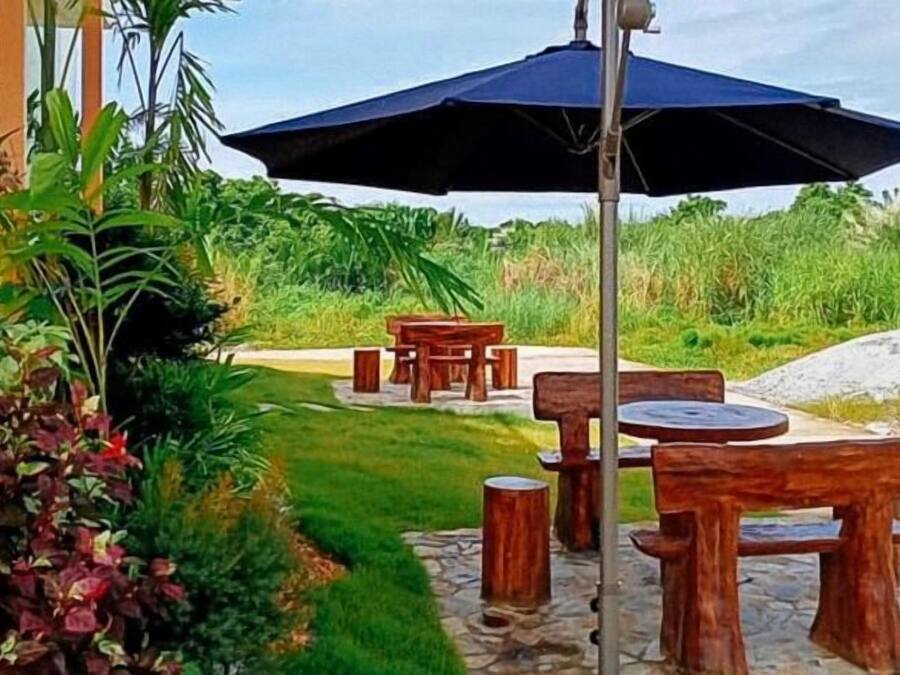 An outdoor seating area with wooden tables and chairs, surrounded by lush greenery and a stone pathway.