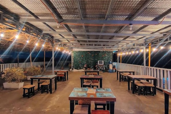 An outdoor dining area with tables and chairs, a green wall with a TV, and string lights.