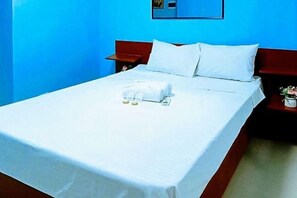 A neatly made bed with white linens and a folded towel, a wooden headboard, and a nightstand with a lamp and a small plant. - Jeanines Travellers Inn by RedDoorz (Morong)