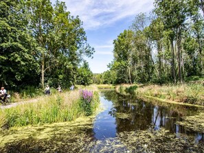 A calm river with dense trees on both sides, a narrow path on the left, and a few people enjoying the outdoors. - Modern Chalet With Dishwasher (Nieuwerkerk aan den IJssel)