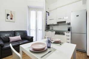 Une cuisine moderne avec des meubles blancs, un réfrigérateur en acier inoxydable et un coin repas comprenant une table blanche et des assiettes roses.