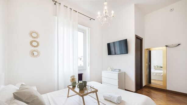 Un dormitorio con cama, mesita de noche con bandeja, televisor, espejo y ventana con cortinas.