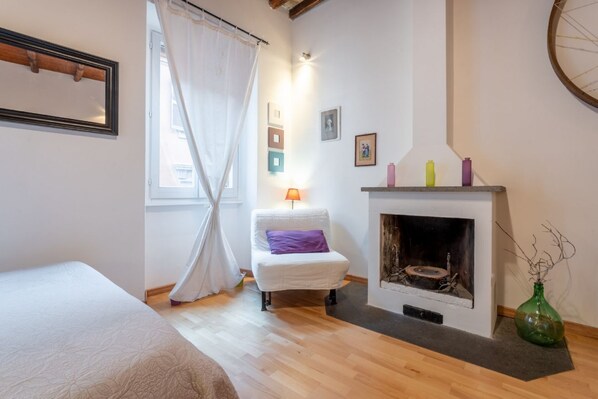 A bedroom with a bed, a white chair, a fireplace, and a window with curtains.