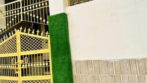 Una reja de metal amarillo con diseños intrincados, un tapete de césped artificial verde y una pared blanca con un pequeño adorno.