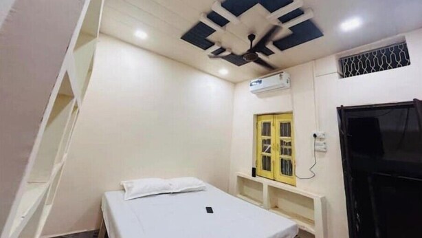 Una habitación pequeña con cama individual, una cama blanca, ventilador de techo, equipo de aire acondicionado, una puerta amarilla y una puerta negra.