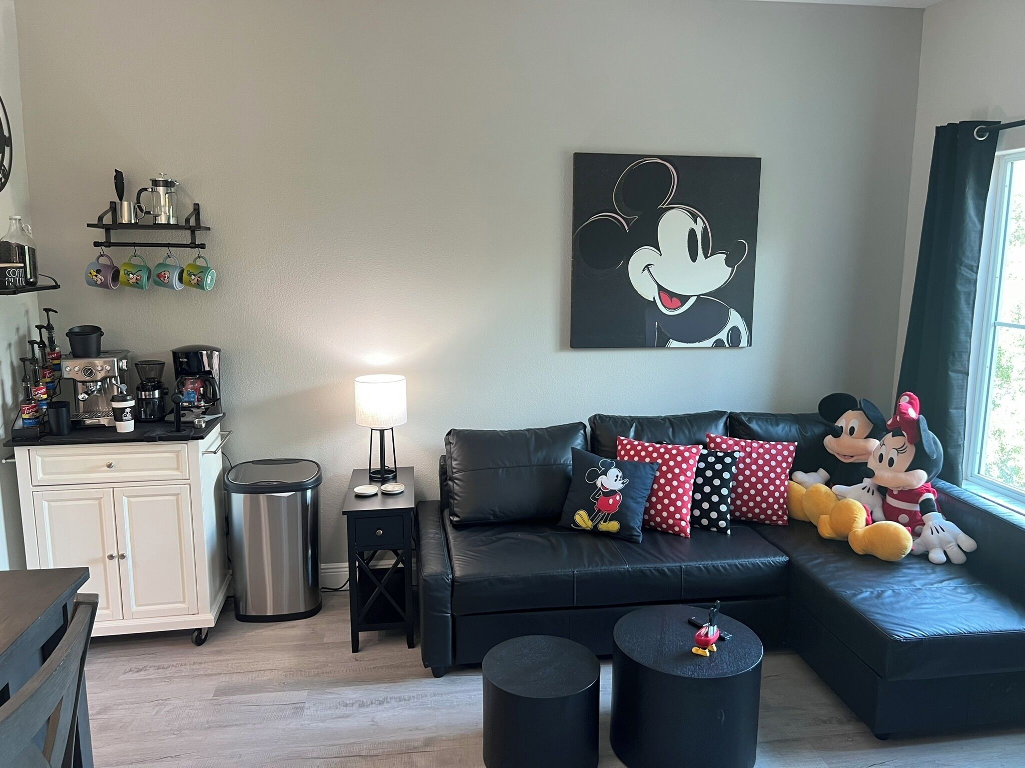 Ein gemütlicher Wohnbereich mit einem schwarzen Sofa, Mickey Mouse Kissen und Plüschspielzeug. Im Hintergrund ist eine Kaffeestation mit verschiedenen Geräten zu sehen.