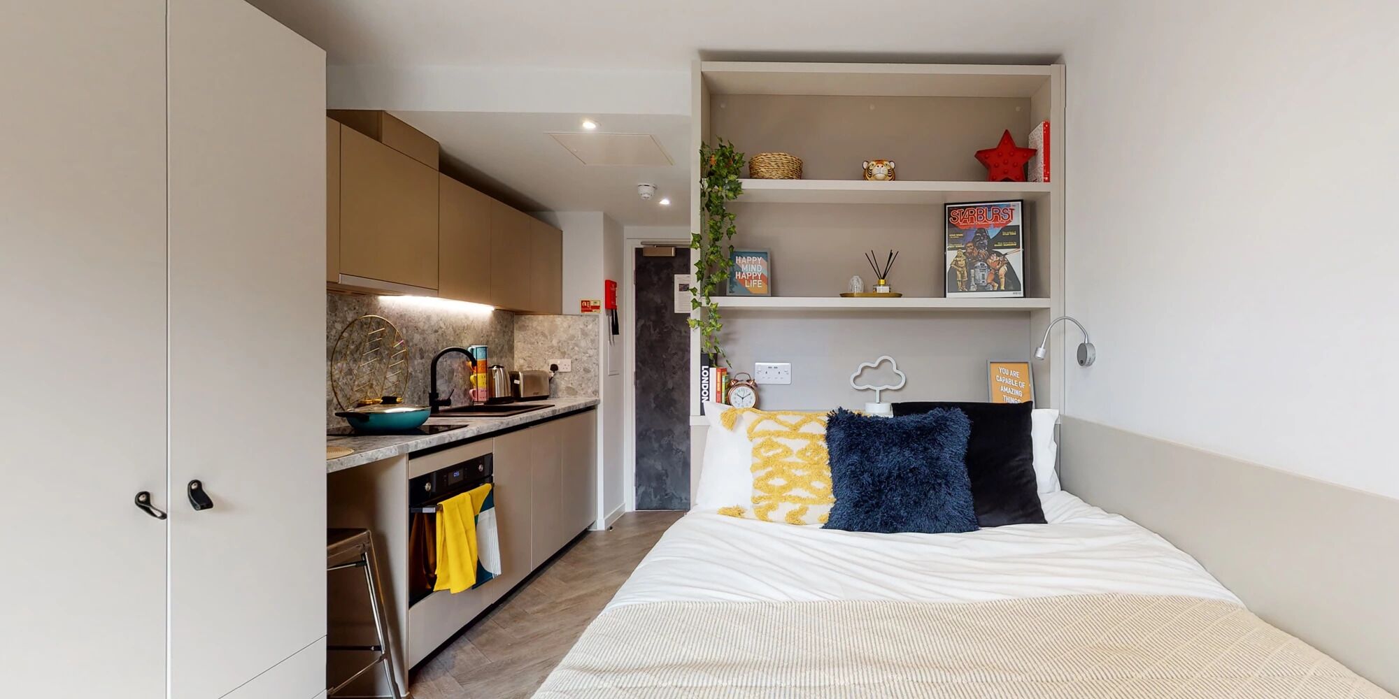 Un espacio habitable compacto con kitchenette, una cama con almohadones decorativos y una estantería con diversos objetos.
