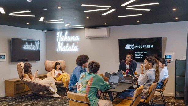 Une salle de réunion moderne dotée d’un grand écran affichant les logos « Hahuna Matata » et « Kissfresh », un groupe de personnes assises autour d’une table avec des ordinateurs portables, et un tapis à motifs.