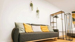 A modern living room with a grey sofa, wooden floor, and minimalist decor including hanging plants and a black metal shelf unit. - Staylight City-Loft | private parking | trade fair (Hannover)