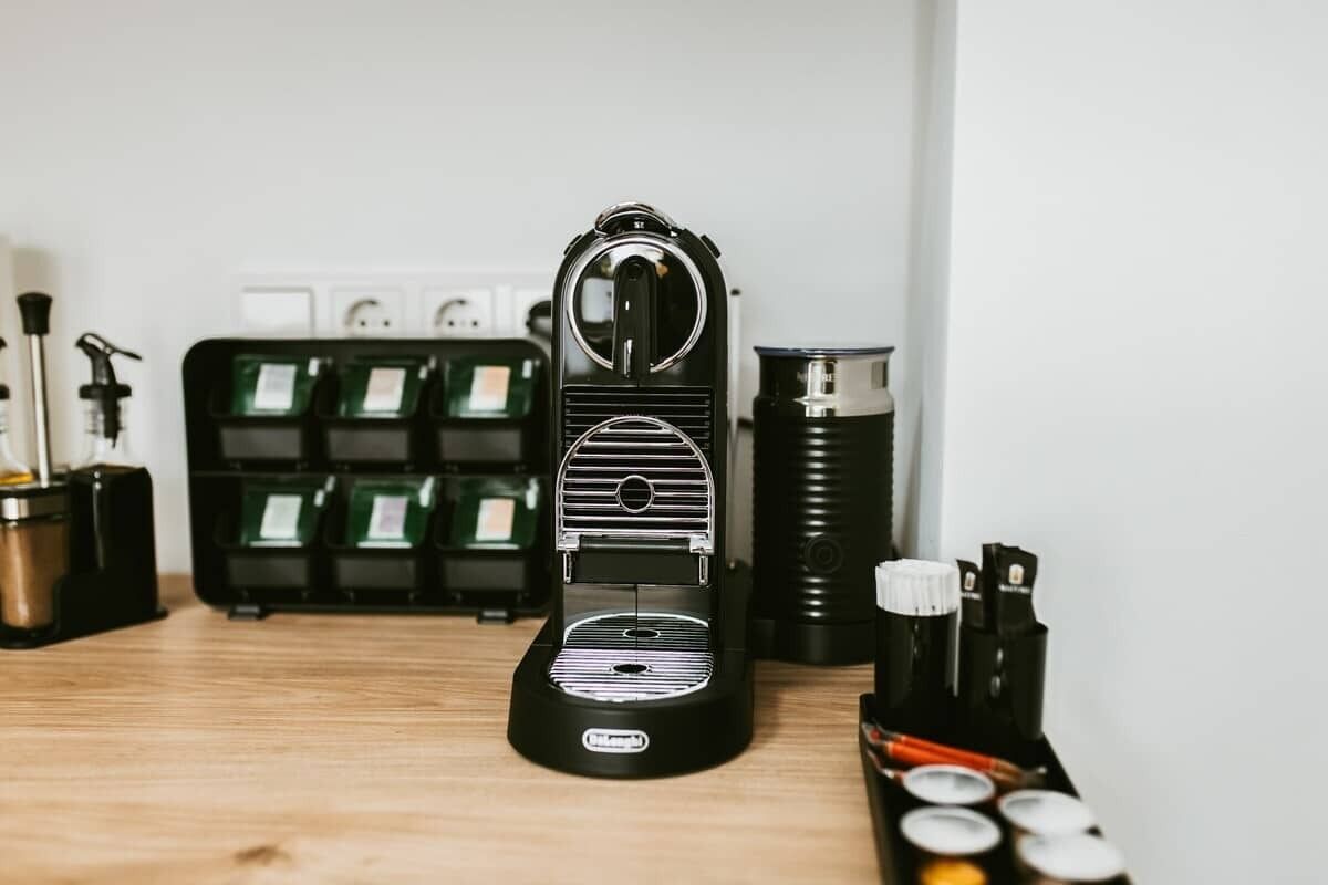A coffee machine on a wooden table with a green basket of coffee pods and a black container of coffee capsules.