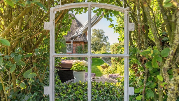 Une maison rustique avec un toit de chaume, entourée d’une végétation luxuriante et dotée d’un cadre de fenêtre blanc.