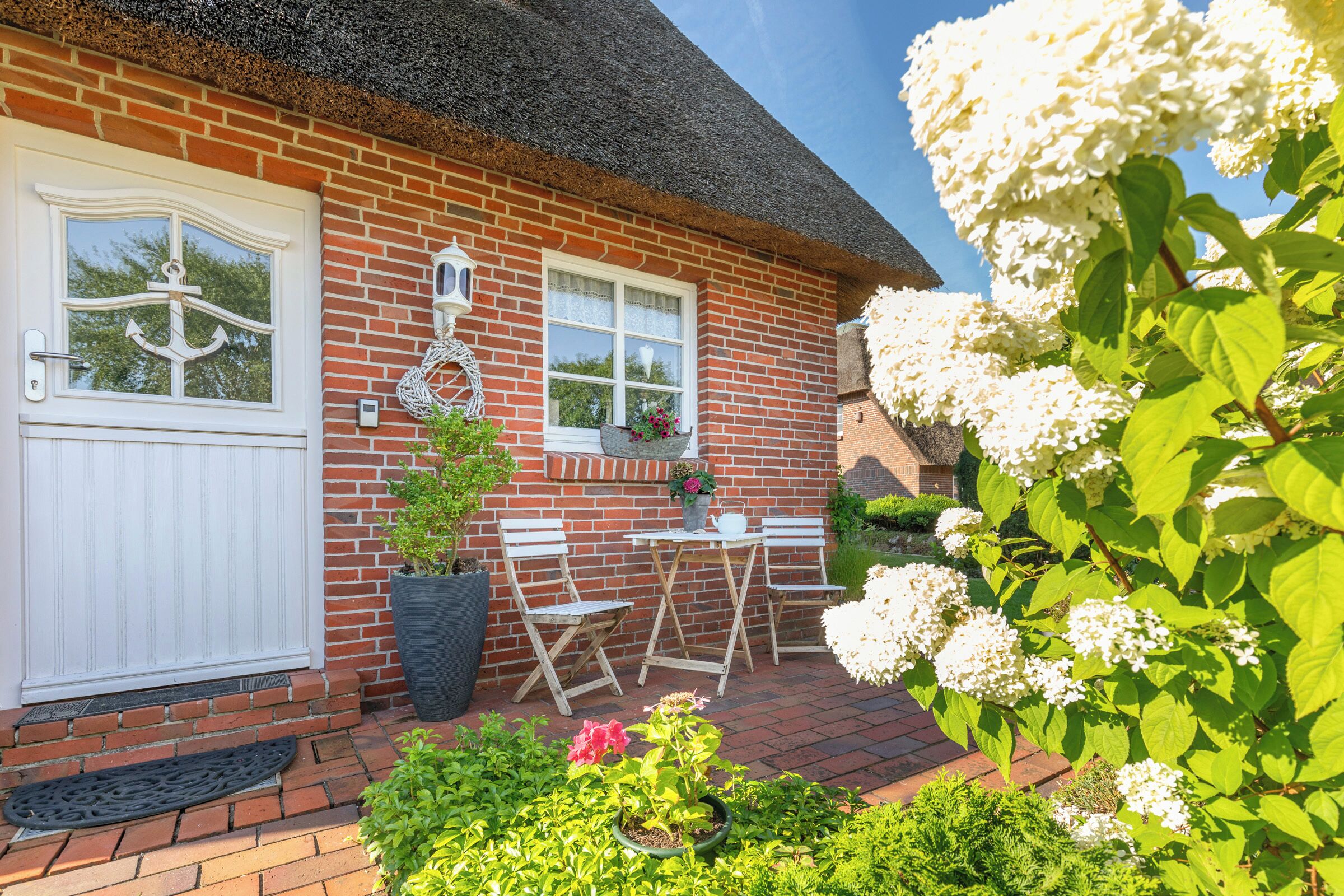 A brick house with a white door, two windows, a thatched roof, and a small patio with a table and chairs.