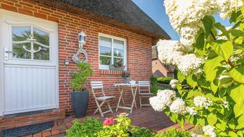 A brick house with a white door, two windows, a thatched roof, and a small patio with a table and chairs.