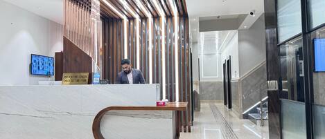 A modern hotel reception area with a marble reception desk, a digital clock, and a check-in/out sign.