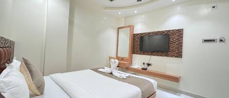 A modern hotel room with a bed, a ceiling fan, a TV mounted on the wall, and a small bedside table with a lamp and a bottle.