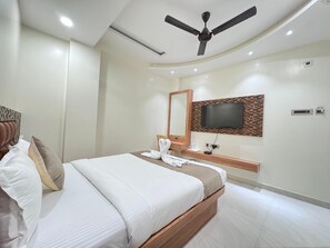 A modern hotel room with a bed, a ceiling fan, a TV mounted on the wall, and a small bedside table with a lamp and a bottle. - HOTEL JIA INTERNATIONAL (Patna)