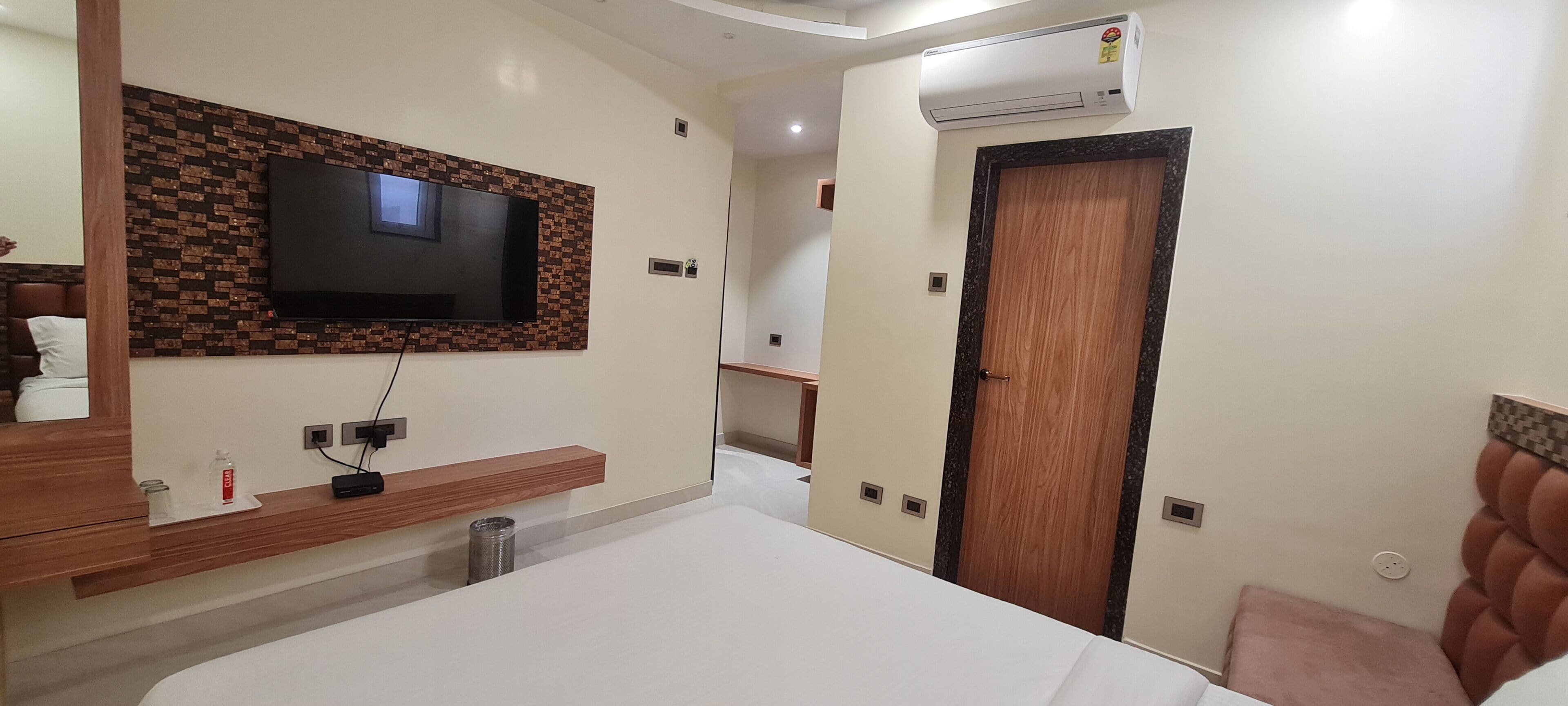 Une chambre d’hôtel moderne avec un téléviseur à écran plat fixé à un mur dont le motif rappelle celui d’un mur de briques, une table de chevet en bois, un lit avec une tête de lit blanche et une porte en bois.