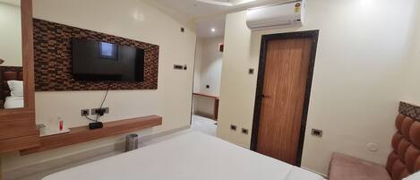 A modern hotel room with a flat-screen TV mounted on a brick-patterned wall, a wooden bedside table, a bed with a white headboard, and a wooden door.