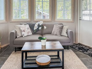A living room with a grey sofa, white pillows, a small table with a plant, and a window with blinds. - Villa taikavuosseli by Interhome (Kuusamo)
