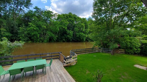 A wooden deck with green chairs overlooking a river, surrounded by lush greenery.
