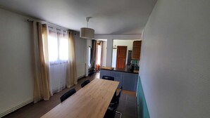 A dining area with a wooden table and chairs, a kitchen in the background, and a window with curtains.