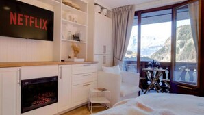 A modern hotel room with a fireplace, a bed, a balcony with a mountain view, and a shelf with books and decorative items. - Stylishly renovated, king bed, 120m from telecabin (Les Houches)