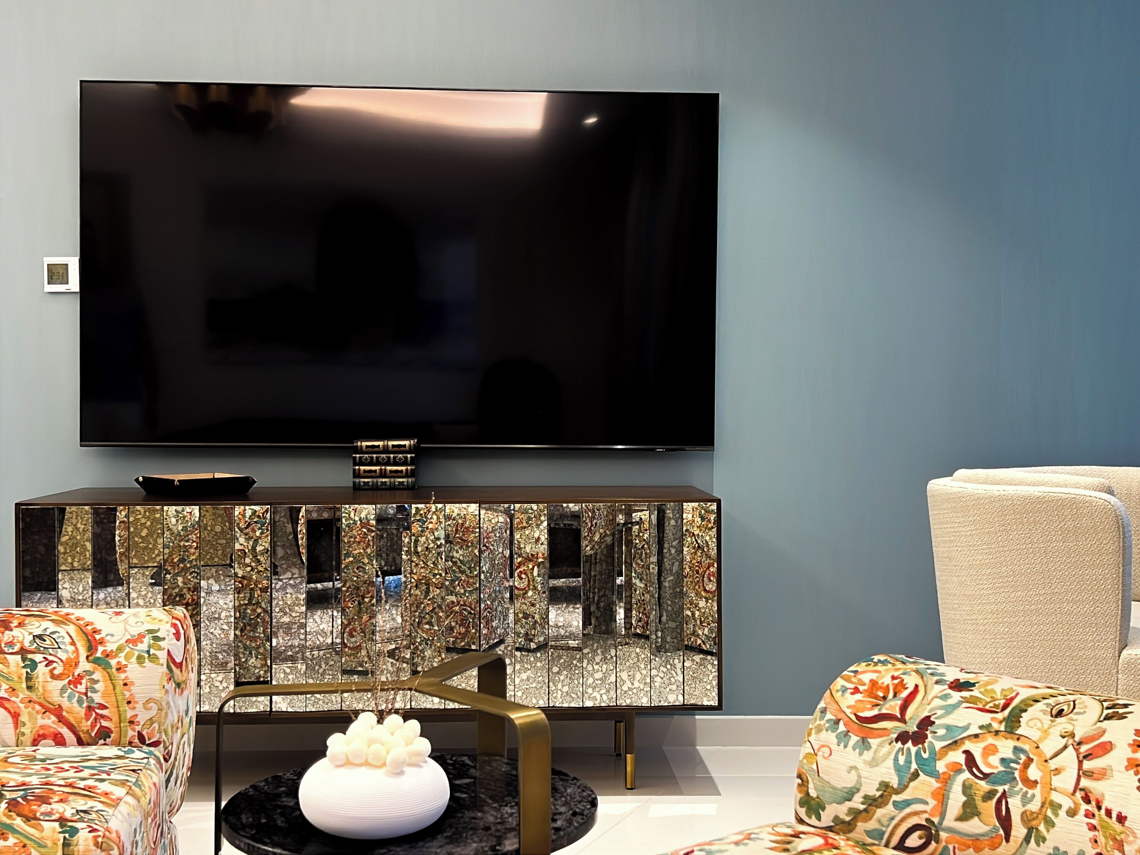 A modern living room with a large flat-screen TV mounted on the wall, a decorative console table with a reflective surface, and two patterned armchairs.
