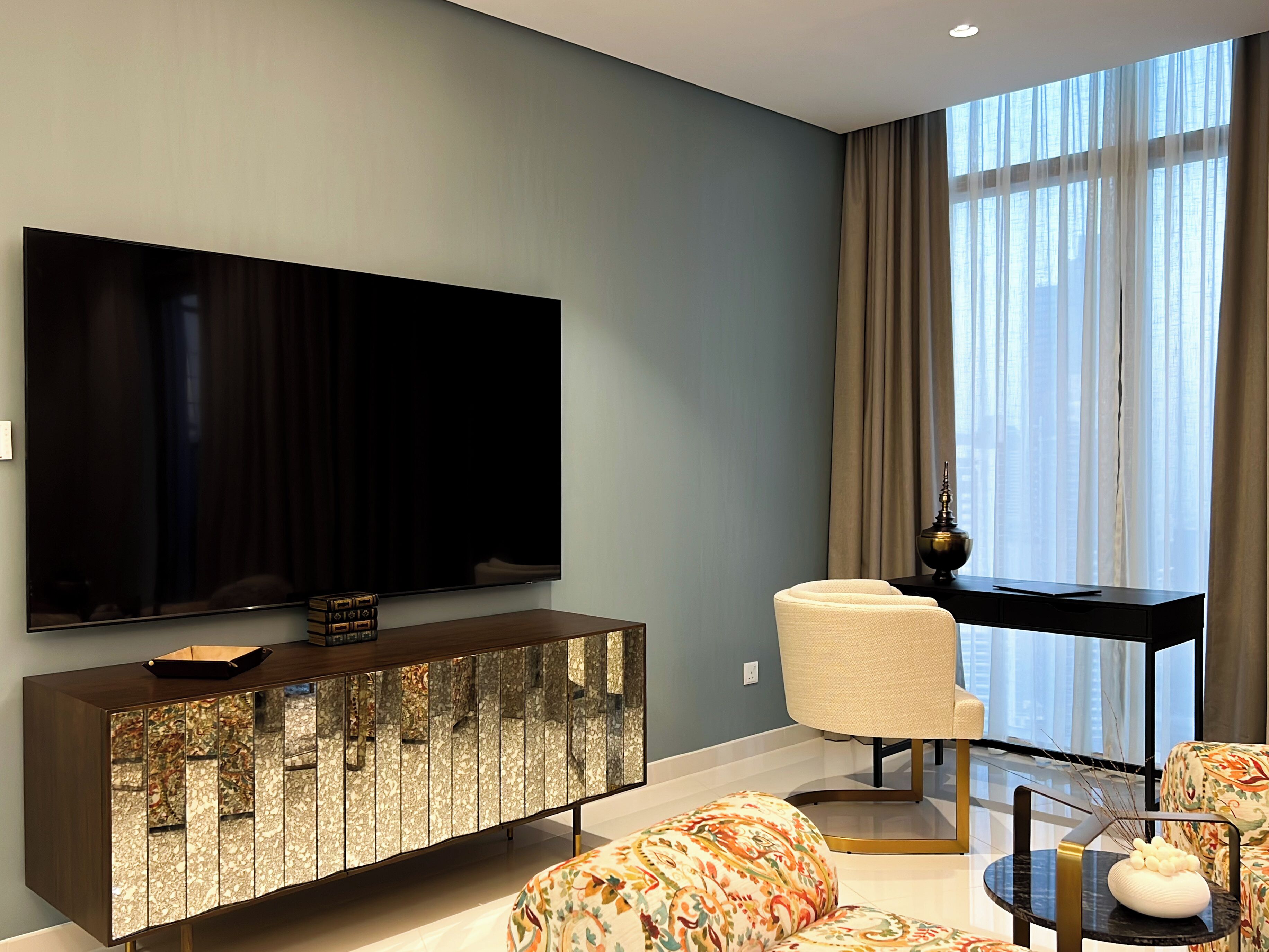 A modern living room with a large flat-screen TV mounted on the wall, a wooden cabinet with a glass front, a white armchair, and a round coffee table with a vase.
