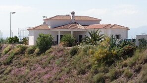 A white building with a tiled roof, surrounded by a landscaped garden with various plants and flowers.