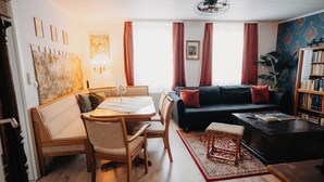 Un salon chaleureux avec une table à manger en bois, des chaises rembourrées, un canapé sombre agrémenté de coussins rouges, une bibliothèque et un grand tableau abstrait accroché au mur.