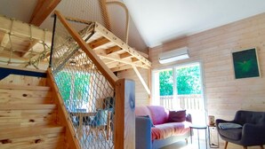 A wooden cabin interior with a staircase, a sofa, a chair, and a window with a view of greenery.