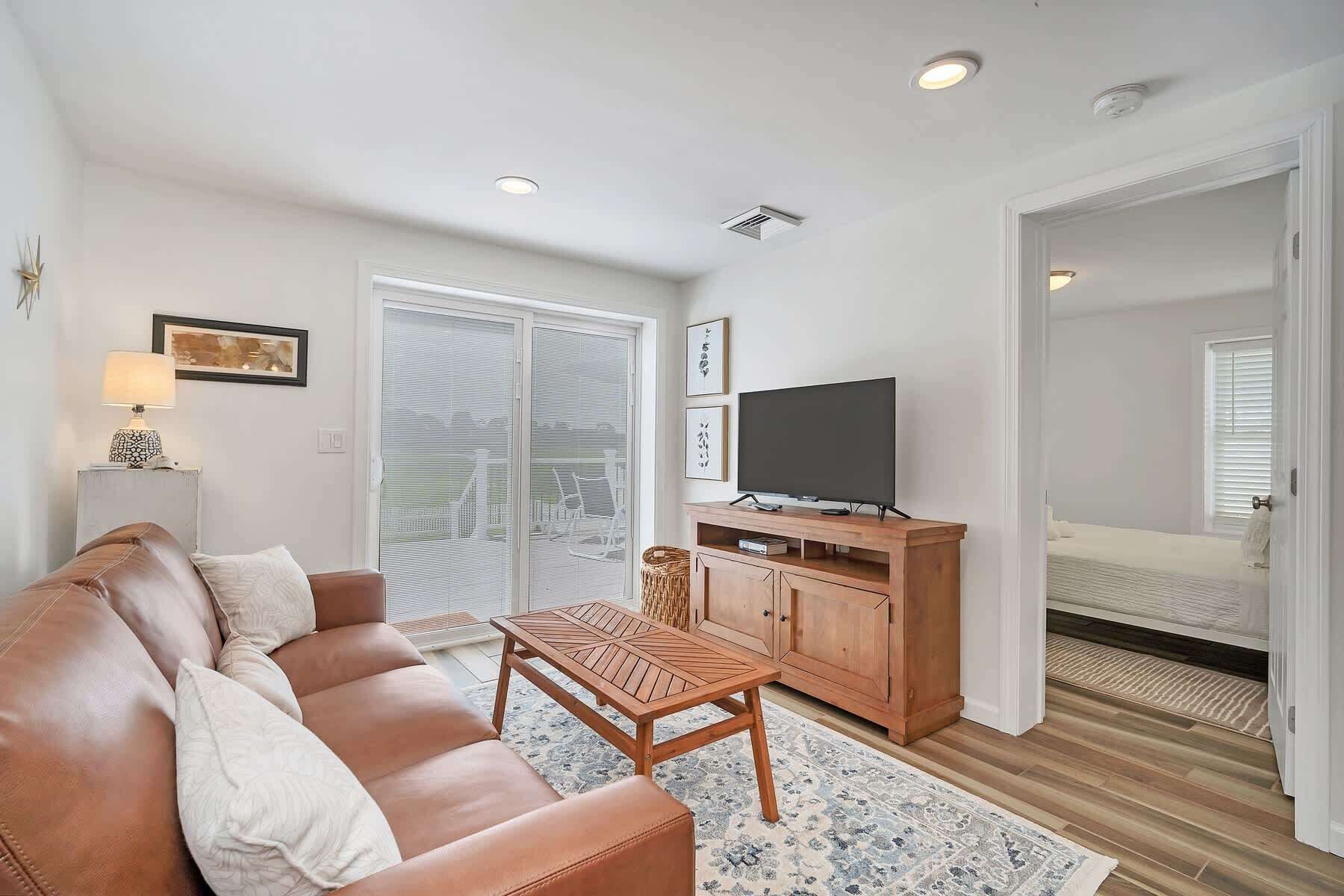 A living room with a sofa, a TV on a wooden stand, a patterned rug, and a sliding glass door leading to a bedroom.