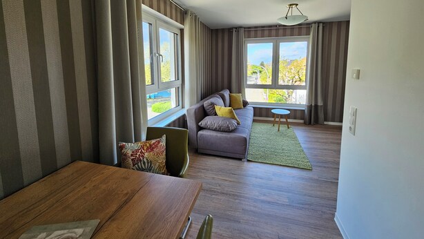 Un salon moderne avec un canapé, une table à manger et une vue sur l’extérieur grâce à de grandes fenêtres.
