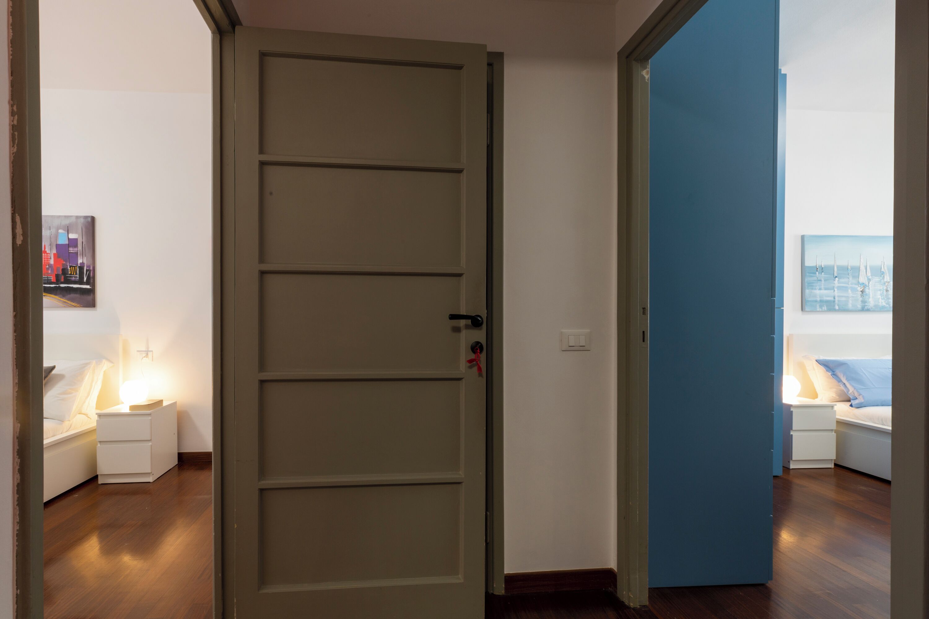 A hotel room with a wooden floor, a bed with a white headboard, a bedside table with a lamp, and a painting on the wall.