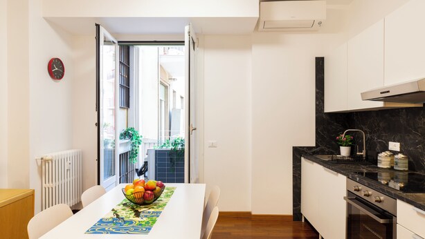 A modern kitchen with a dining area, featuring a white table with a fruit bowl, a clock on the wall, and a view through an open door. - notaMi - Rainbow Home - CityLife (Milan)