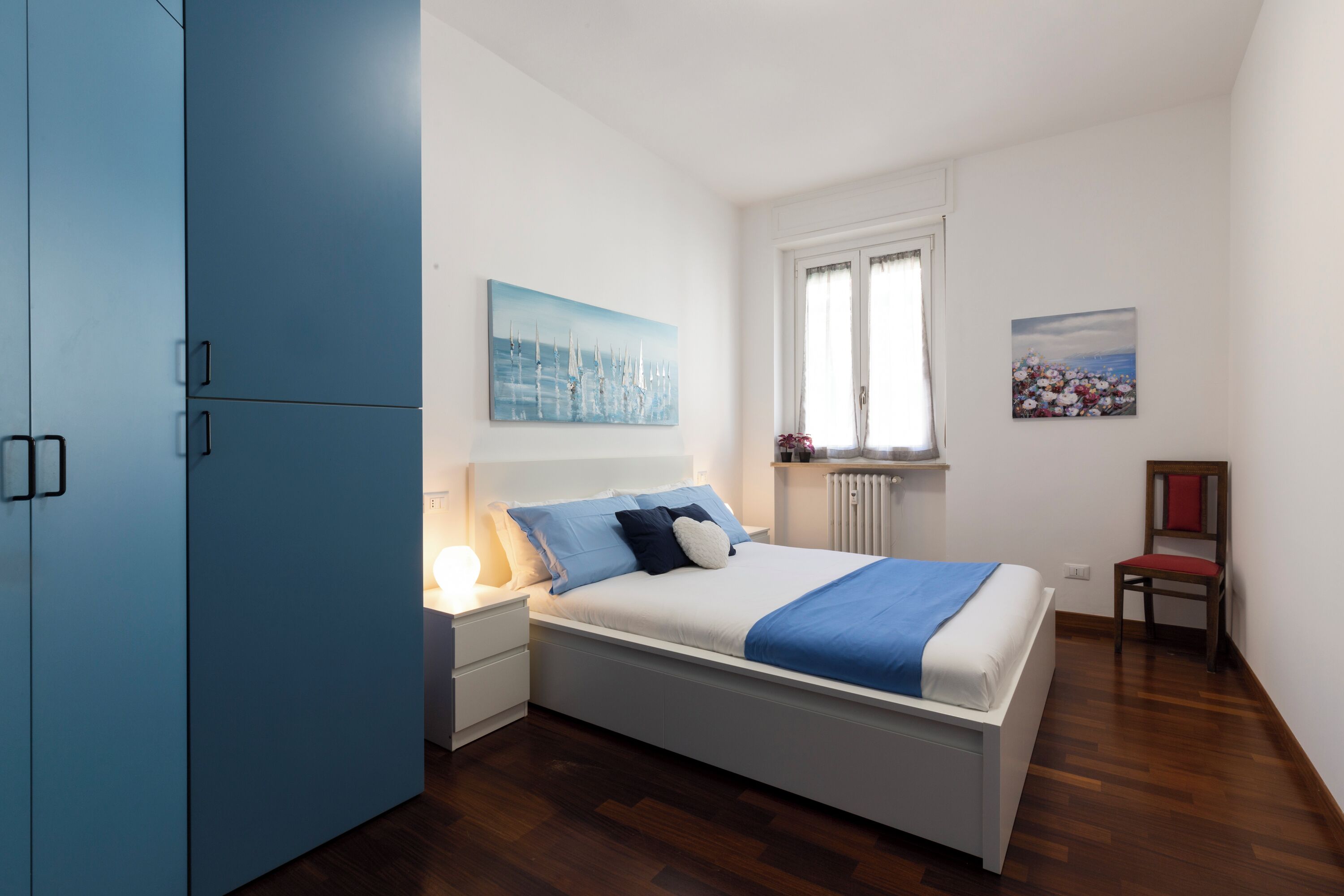 A bedroom with a bed, nightstands, a blue wardrobe, and artwork on the walls.